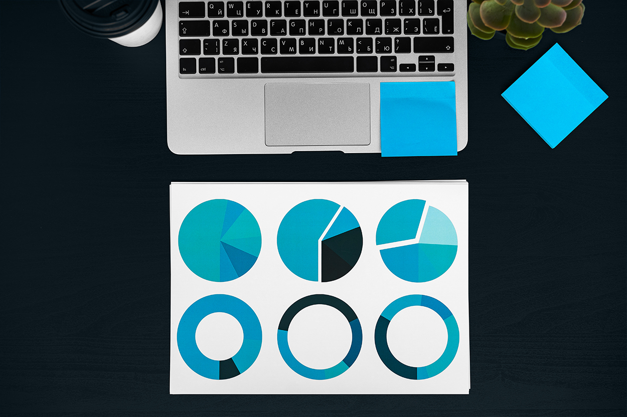 Laptop-Tastatur, darunter ein Papier mit Diagrammen, daneben Post-its und ein Kaffeebecher auf dunklem Schreibtisch.