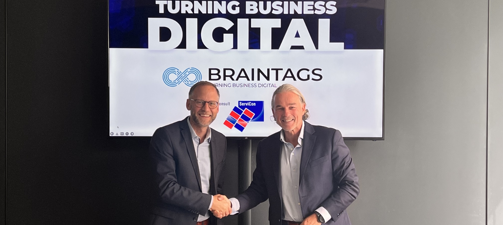 Servicon- und Braintags-Geschäftsführer schütteln sich die Hand vor einem Bildschirm mit Turning Business Digital Schriftzug